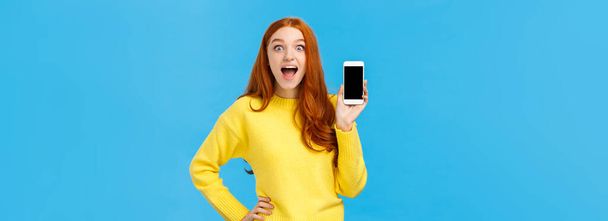 Lenyűgözött és lenyűgözött, izgatott vörös hajú nő sárga pulóverben bemutatja az új alkalmazást, bemutatja okostelefon kijelző, mosolygós lenyűgözött nyitott száj szórakozott, néz kamera, kék háttér. - Fotó, kép