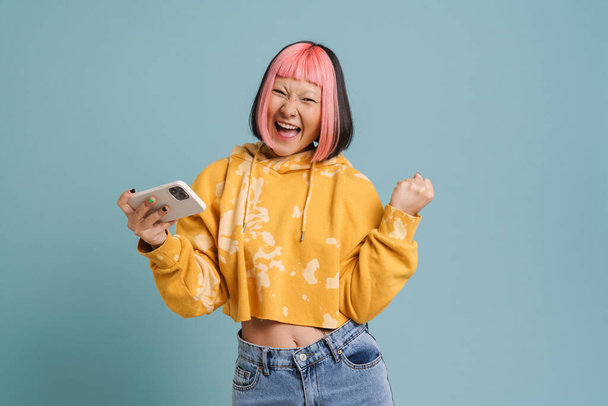 Mavi arka planda izole edilmiş cep telefonuyla çevrimiçi oyun oynarken el kol hareketi yapan Asyalı kız - Fotoğraf, Görsel