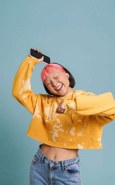 Asian girl in earphones dancing while listening music with cellphone isolated over blue background - Foto, immagini