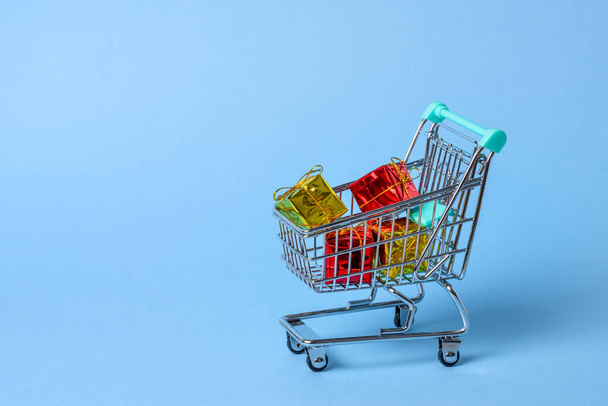 Close up of a small shopping cart full of colourful Christmas gifts on blue background. Copy space. - Photo, Image