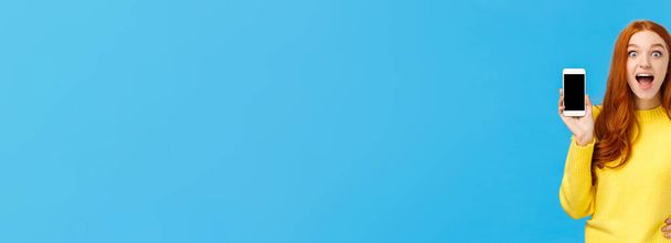 Sorprendido e impresionado, excitado pelirroja hembra en suéter amarillo introducir nueva aplicación, mostrando la pantalla del teléfono inteligente, sonriendo fascinado boca abierta divertido, mira la cámara, fondo azul
. - Foto, Imagen
