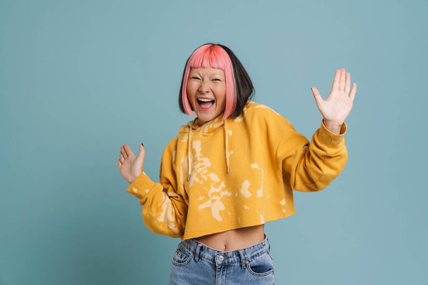 Азиатская девушка с розовыми волосами и пирсинг выражая удивление перед камерой изолированы на синем фоне - Фото, изображение