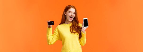 Shopping, Werbung und mobiles Suchtkonzept. attraktive freche rothaarige Mädchen zeigt Smartphone und Kreditkarte, zwinkert und lächelt Kamera, fördert Online-Banking, Einzahlungs- oder Zahlungsmethode. - Foto, Bild