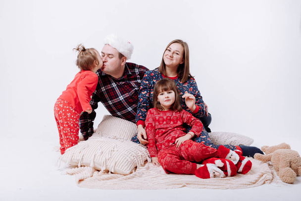 Молодая счастливая европейская семья в рождественской пижаме: улыбающиеся мама, папа и две маленькие дочери сидят на теплом пледе на белом фоне. Тема семейного праздника - Новый год и Рождество - Фото, изображение