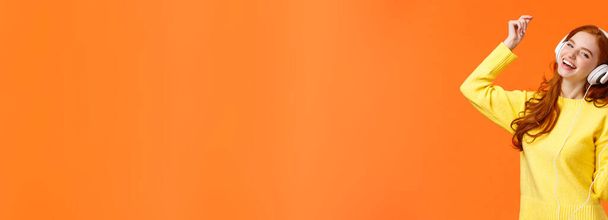 Adolescente rousse heureuse insouciante reçoit de nouveaux écouteurs pour cadeau de Noël, danse lever la main tout en écoutant de la musique préférée, tenant smartphone, mettre une piste impressionnante, fond orange
. - Photo, image
