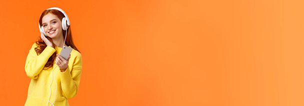 Waist-up portret vrolijke mooie jonge vrouw met rood haar luisteren muziek in hoofdtelefoon, kantelen hoofd en glimlachen tevreden, holding smartphone, kies lied voor zachte rustige avond, oranje achtergrond. - Foto, afbeelding