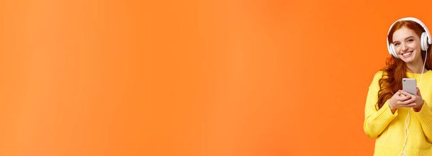 Technologie, Shopping und Menschen-Konzept. modernes nettes Hipster-Rotschopf-Mädchen mit weißen Kopfhörern, Smartphone in der Hand, unbeschwert lächeln, angenehmen Musiksound genießen, orangefarbener Hintergrund im Stehen. - Foto, Bild