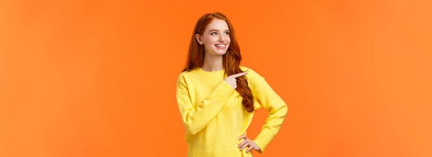 夢のような、活気に満ちた良い黄色のセーターの赤毛の女性は、優れた休日の贈り物を発見し、消費者の左を指して、満足して見て、ピッキング製品として笑みを浮かべて、意思決定、オレンジの背景. - 写真・画像