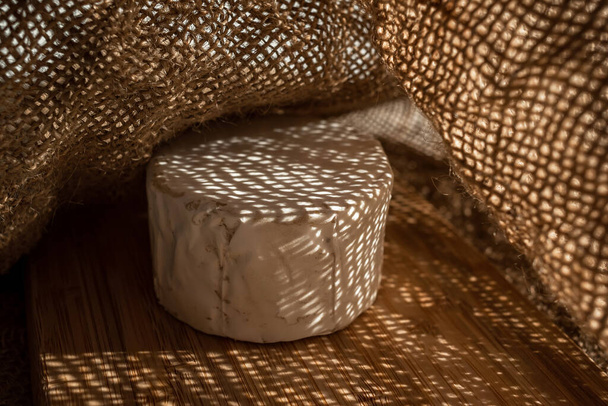 A composition of a round cheese head with white mold on a bamboo board on a background of burlap. Aesthetic serving of food - Фото, зображення