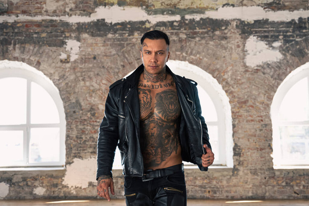 hot tattooed man in leather against the big window.Handsome Young Athletic Male Fashion Model.Muscular athletic sexy male with naked torso. Brutal handsome man with tattooed body. - Photo, image