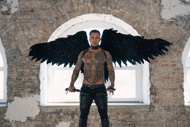 翼を持つハンサムな男黒い翼を持つ男性天使。鞭で筋肉の無気力な男。入れ墨の体を持つ残忍なハンサムな男。筋肉運動セクシー男性  - 写真・画像