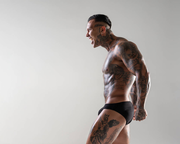 Jonge knappe bodybuilder poseren, atleet toont bodybuilding poseren. hete getatoeëerde man met mooi lichaam toont zijn spieren in de studio - Foto, afbeelding