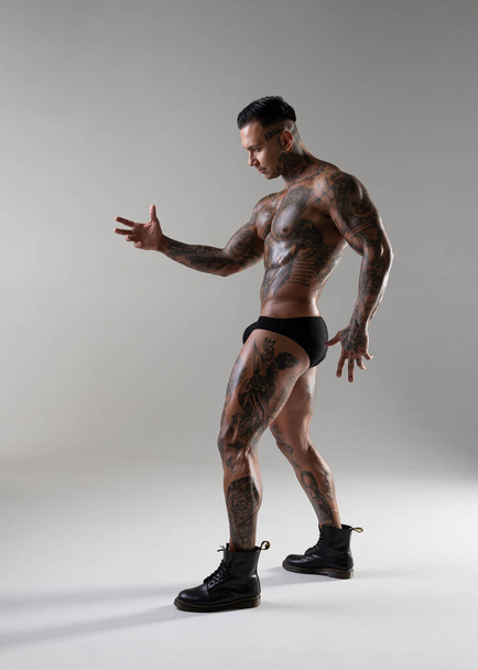 Молодий красивий культурист позує, спортсмен показує позування бодібілдингу. гарячий татуйований чоловік з красивим тілом показує свої м'язи в студії
 - Фото, зображення