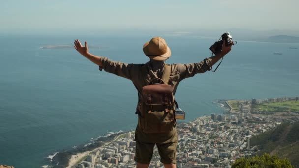 climber standing on the top of a tall mountain looking at and admiring the panorama sunset. male with a backpack stands on the edge of a cliff near the ocean. Man raising hands on top of the mountain - Footage, Video