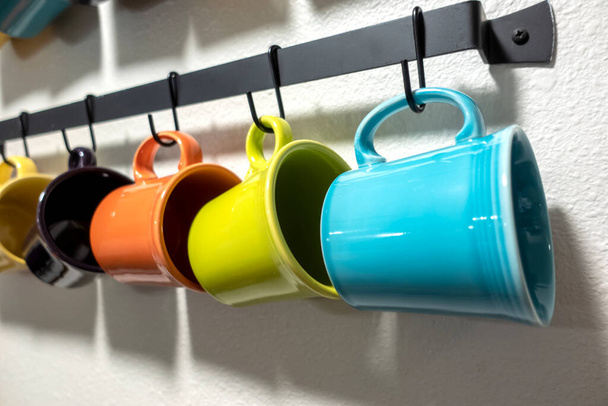 A collection of colorful mugs hanging on black hooks against a white wall - Фото, изображение
