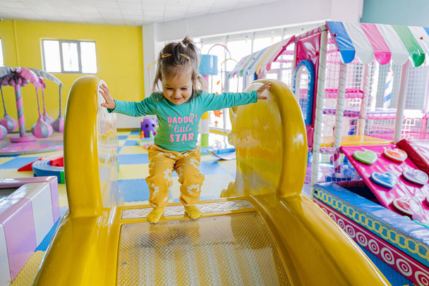 Happy girl playing at indoor play center playground. Sliding in yellow slide. - Zdjęcie, obraz