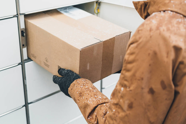 A man with a box in his hands near the self-service mail terminal. Parcel delivery machine. Person holding a cardboard box. Mail delivery and post service, online shopping, e commerce concept - Photo, Image