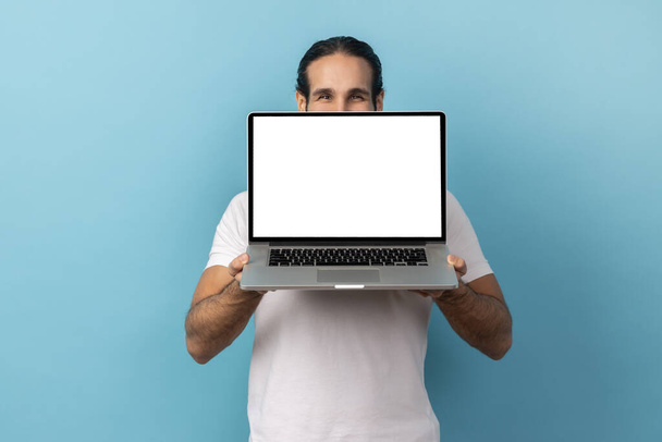 Portrait of man with beard wearing white T-shirt covering half of face behind laptop with white empty display, looking at camera with kind eyes. Indoor studio shot isolated on blue background. - Foto, Bild