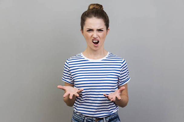 Portrait of angry unhappy frustrated woman wearing striped T-shirt standing spreading hands with shock and misunderstanding, how could you sign. Indoor studio shot isolated on gray background. - Φωτογραφία, εικόνα
