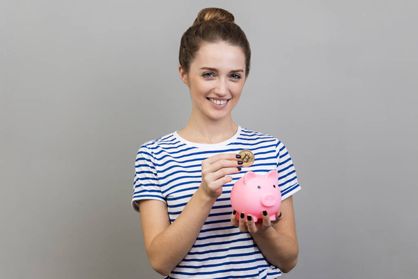 Portrait of woman wearing striped T-shirt putting physical bitcoin into piggy bank, investment in cryptocurrency concept, looking smiling at camera. Indoor studio shot isolated on gray background. - Fotografie, Obrázek