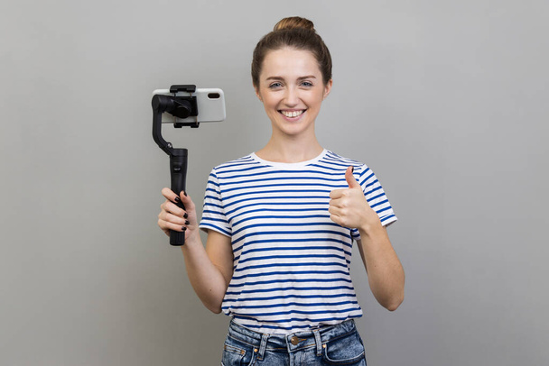 Satisfied stylish woman wearing striped T-shirt holding mobile phone and steadicam, looking at camera with positive expression, showing thumb up. Indoor studio shot isolated on gray background. - Foto, Bild