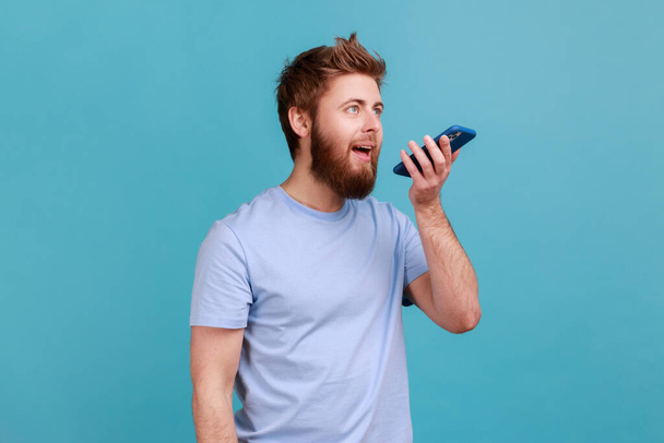 Portrait of positive optimistic bearded man recording voice message or talking to online assistant at his smartphone holding phone near mouth. Indoor studio shot isolated on blue background. - Foto, Imagem