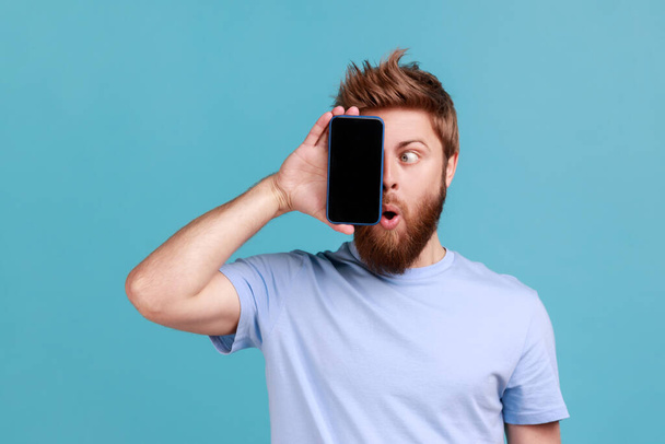 Portrait of bearded man covering eye with cellphone, hiding half of face, using gadget with wow shocked expression, trying to look at display. Indoor studio shot isolated on blue background. - 写真・画像
