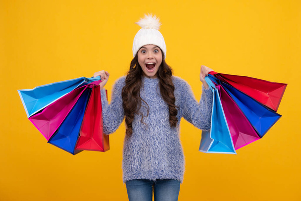 Teenager child girl in autumn hat with shopping bags on yellow background. Kid with shopping winter sale bags. Excited face, cheerful emotions of teenager girl - Fotografie, Obrázek