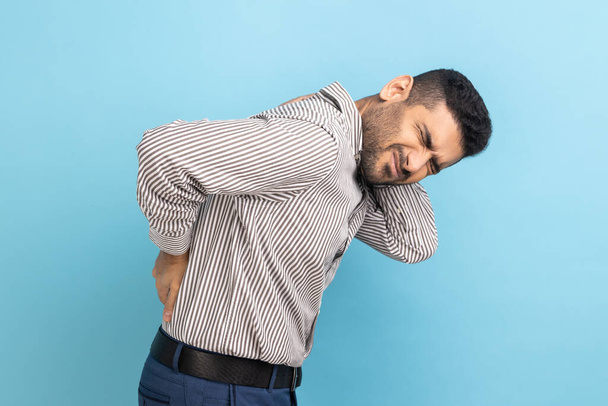 Backache or spine or kidney pain. Portrait of young sick businessman standing and touching his back and feeling pain, wearing striped shirt. Indoor studio shot isolated on blue background. - Foto, Imagem