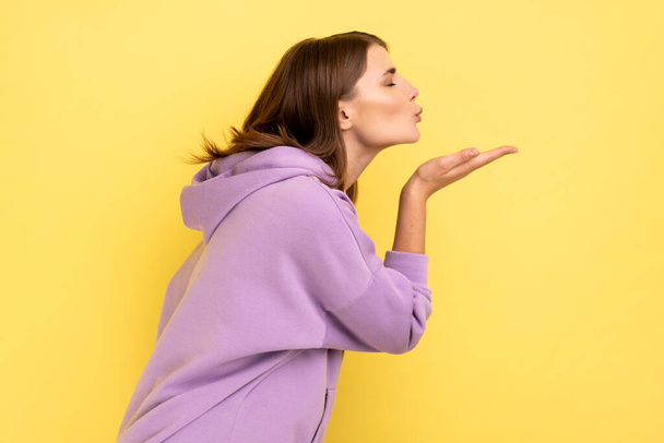 Side view of woman sending air kiss, flirting, demonstrating love affection feelings, keeps eyes closed, romantic relationships, wearing purple hoodie. Indoor studio shot isolated on yellow background - Valokuva, kuva