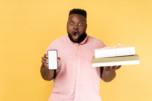 Portrait of shocked astonished man in pink shirt holding present box and smatrphone with blank screen for promotion, looking at camera with big eyes. Indoor studio shot isolated on yellow background. - Foto, Imagen