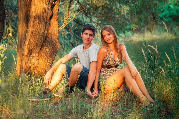 Junge und Mädchen halten Händchen. Der Typ liebt das Mädchen. Junge Verliebte sitzen am Ufer des Sees in der Nähe des Baumes. - Foto, Bild