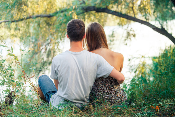 Junge und Mädchen halten Händchen. Der Typ liebt das Mädchen. Junge Verliebte sitzen am Ufer des Sees in der Nähe des Baumes. - Foto, Bild