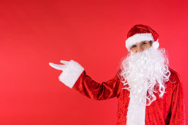 Man dressed as Santa Claus, pointing to the side with his finger, on red background. Christmas, celebration, gifts, consumerism and happiness concept. - Photo, Image