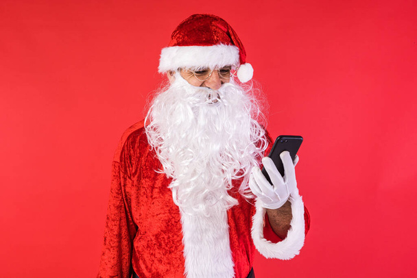 Чоловік, одягнений як Санта Клаус, консультується зі своїм мобільним телефоном на червоному тлі. Різдво, свято, подарунки, споживання і щастя.. - Фото, зображення