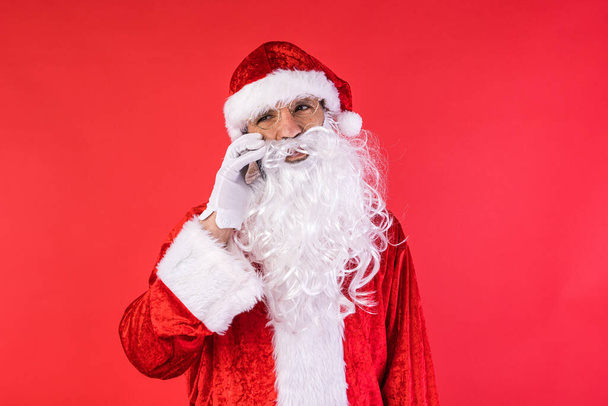 Homme habillé comme le Père Noël, parlant sur son téléphone portable, sur fond rouge. Noël, célébration, cadeaux, concept de consumérisme et de bonheur. - Photo, image