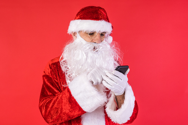 Człowiek przebrany za Świętego Mikołaja, sprawdzający komórkę, na czerwonym tle. Boże Narodzenie, świętowanie, prezenty, konsumpcjonizm i koncepcja szczęścia. - Zdjęcie, obraz