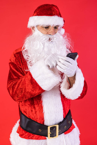 Muž oblečený jako Santa Claus, konzultující svůj mobil, na červeném pozadí. Vánoce, oslavy, dárky, konzumerismus a koncept štěstí. - Fotografie, Obrázek