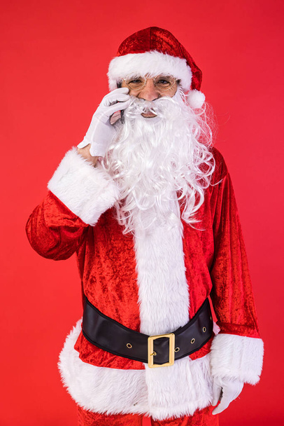 男はサンタクロースの格好をし、携帯電話で赤い背景で話していた。クリスマス、お祝い、贈り物、消費主義と幸福の概念. - 写真・画像