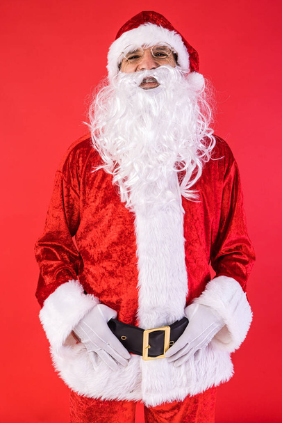 Kırmızı arka planda kameraya poz veren Noel Baba kılığındaki bir adamın portresi. Noel, kutlama, hediyeler, tüketim ve mutluluk kavramı. - Fotoğraf, Görsel
