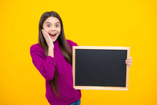 Teenager younf school girl holding school empty blackboard isolated on yellow background. Portrait of a teen female student. Excited face. Amazed expression, cheerful and glad - Photo, Image