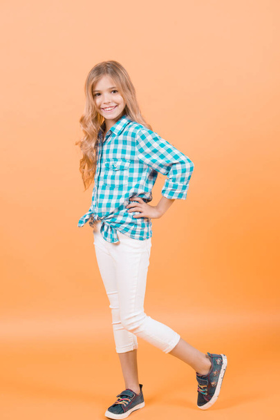 Child model smile with long blond hair. Girl in plaid shirt, pants, sneakers on orange background. Kid fashion, style, trend. Beauty, look, hairstyle. Happy childhood concept. - Foto, afbeelding