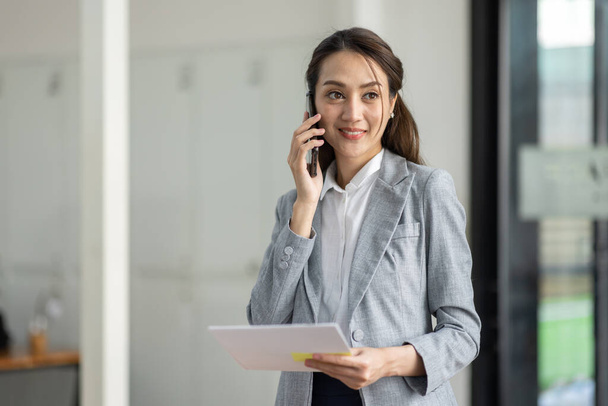 Jeune femme asiatique souriante d'affaires parlant sur le téléphone mobile, debout dans le bureau de travail avec le document dans sa main - Photo, image