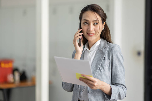 Молодая улыбающаяся деловая азиатка разговаривает по мобильному телефону, стоя в офисе на рабочем месте с документом в руке - Фото, изображение