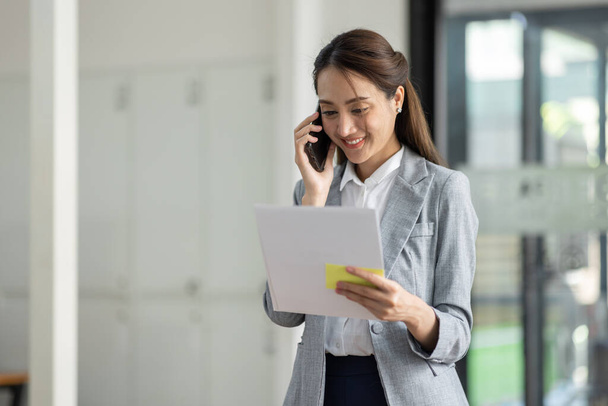 Fiatal mosolygós Üzleti ázsiai nő beszél mobiltelefonon, áll a munkahelyi irodában dokumentummal a kezében - Fotó, kép