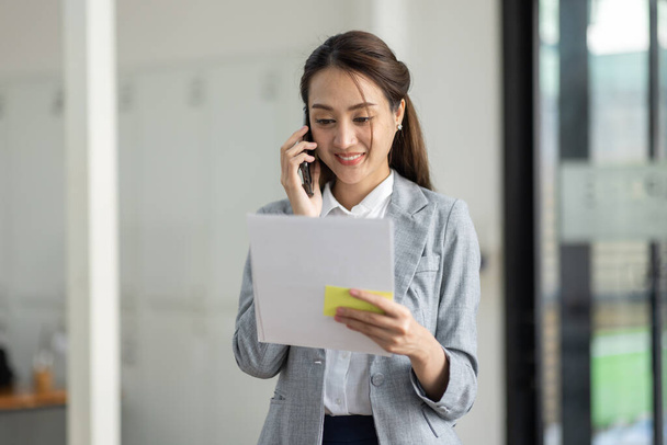 Mladá usměvavá asijská žena mluví na mobilním telefonu, stojí v kanceláři na pracovišti s dokumentem v ruce - Fotografie, Obrázek