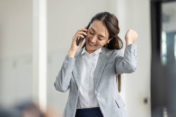 Молодая улыбающаяся счастливая деловая азиатка разговаривает по мобильному телефону, стоя в офисе на рабочем месте с документом в руке - Фото, изображение