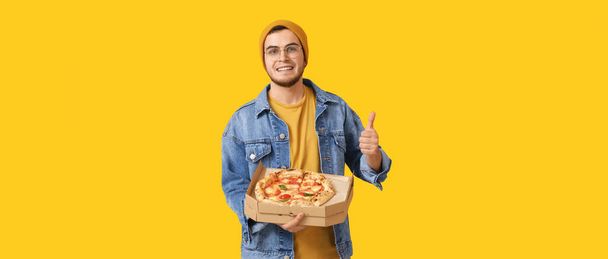 Beau jeune homme avec une délicieuse pizza montrant pouce vers le haut sur fond jaune - Photo, image