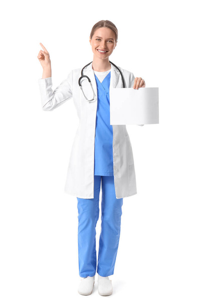 白い背景に何かを指している空白の紙のシートを持つ女性医師 - 写真・画像