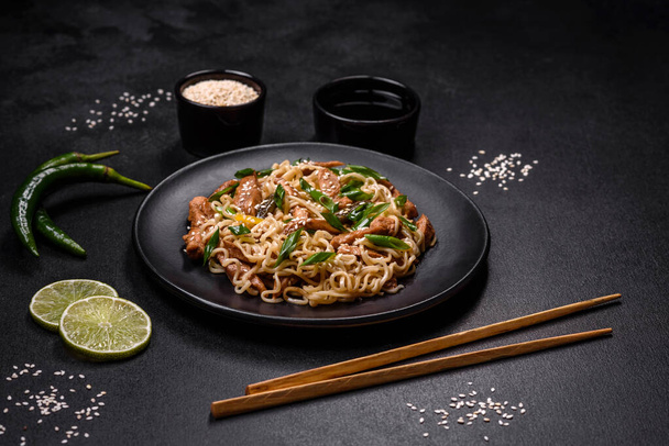 Tasty dish of Asian cuisine with rice noodles, chicken, asparagus, pepper, sesame seeds and soy sauce on dark concrete background - Photo, Image